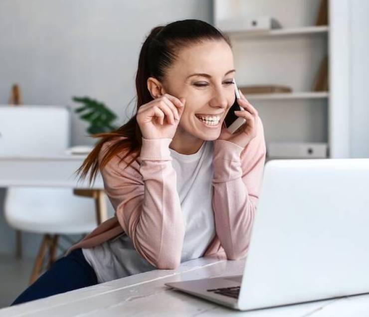 Woman talking by phone while working on laptop indoors